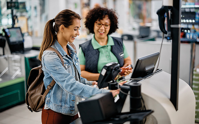 Young happy woman using self-service checkout with help of supermarket worker.