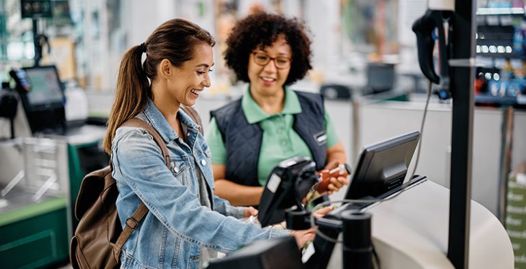 Young happy woman using self-service checkout with help of supermarket worker.